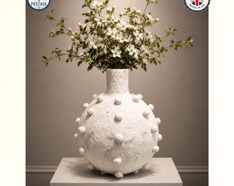 May include: A round, white, textured vase with a narrow neck and small, evenly spaced spherical protrusions across its surface. The vase is filled with a bouquet of delicate white flowers with green leaves and thin, branching stems.