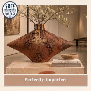 Peut inclure: Un vase en céramique large en forme de losange avec une surface texturée brun foncé rugueuse et des coulures verticales de couleur claire près du col. Le vase contient des branches fines à petites fleurs blanches et deux livres empilés.