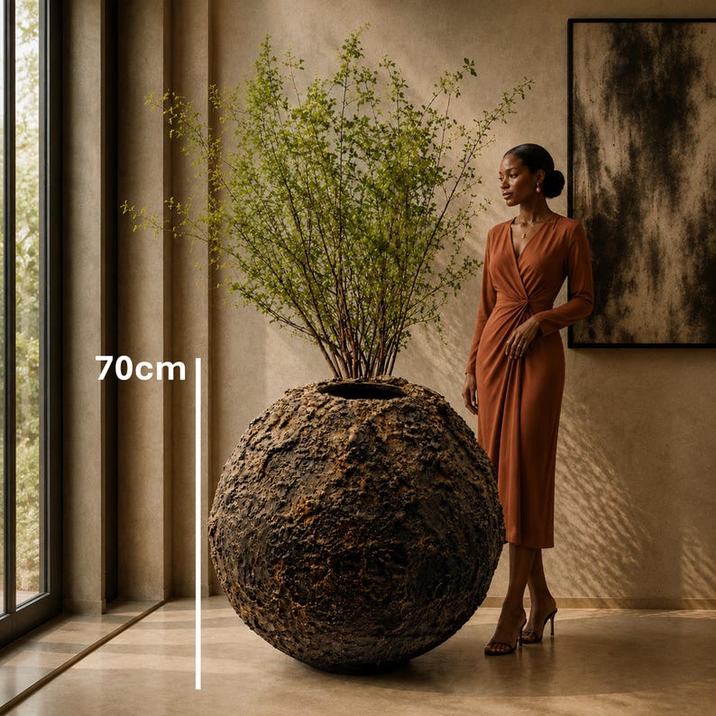 Op de afbeelding: Een grote, bolvormige plantenbak met een donker, zwaar getextureerd en gekraterd oppervlak. De plantenbak heeft een ronde opening aan de bovenkant met een groene plant. Een witte verticale lijn en de tekst 70cm geven een hoogte van 70cm aan.
