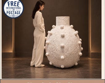 May include: A large, off-white, textured floor vase with a spherical body and a cylindrical neck. The surface of the vase is covered in evenly spaced, protruding spherical bumps. The vase stands on a dark, polished floor surface.