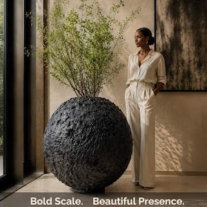 Puede incluir: Una maceta esférica grande de color negro con una superficie rugosa y muy texturizada que contiene una planta verde alta y frondosa. El texto "Bold Scale. Beautiful Presence." está impreso en blanco en la parte inferior.