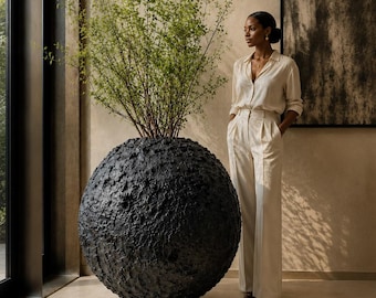 May include: A large, spherical black planter with a heavily textured, rugged surface containing a tall, leafy green plant. The text "Bold Scale. Beautiful Presence." is printed in white at the bottom of the image.