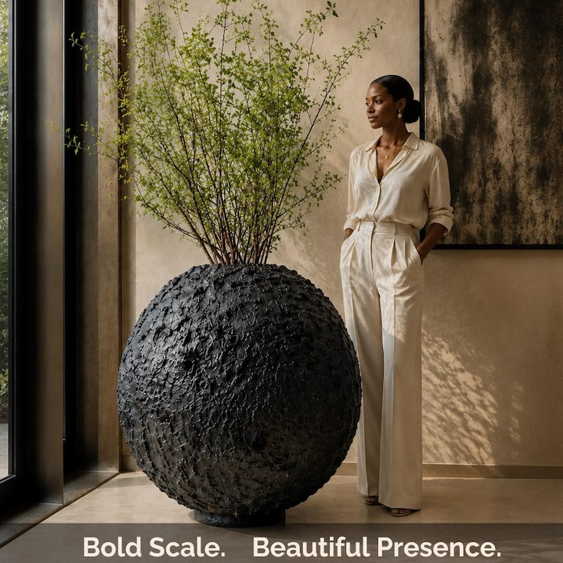 Op de afbeelding: Een grote, bolvormige zwarte plantenbak met een zwaar getextureerd, ruw oppervlak met een hoge, bladerrijke groene plant. De tekst "Bold Scale. Beautiful Presence." staat in het wit onderaan de afbeelding gedrukt.