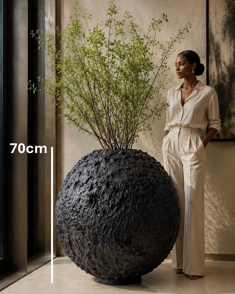 Op de afbeelding: Een grote, bolvormige zwarte plantenbak met een zwaar getextureerd, ruw oppervlak. De plantenbak is voorzien van een zichtbare maataanduiding van 70 cm hoog. Een hoge, bladerrijke groene plant met dunne, vertakte stengels groeit uit de bovenkant.