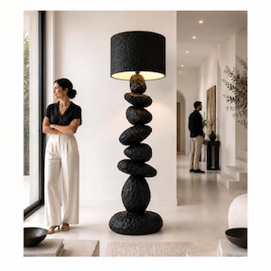 Op de afbeelding: Een vloerlamp met een stapel van zeven zwarte, getextureerde, steenachtige vormen die de voet en de centrale kolom vormen, bekroond met een cilindrische zwarte lampenkap die warm licht uitstraalt vanuit de binnenkant.