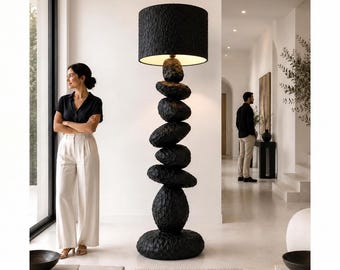May include: A floor lamp featuring a stack of seven black, textured, stone-like shapes forming the base and central column, topped with a cylindrical black lampshade that emits a warm light from the interior.