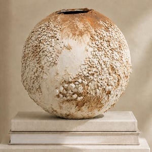 May include: A spherical vase with a textured, off-white and tan surface featuring small, embedded stone-like particles. The vase has a circular opening at the top and a rough, uneven finish across its rounded body, creating a tactile appearance.