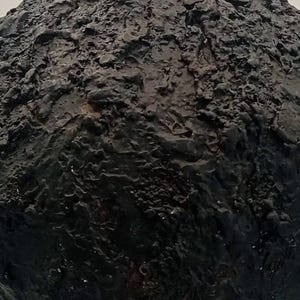 Op de afbeelding: Een bolvormig object met een donkerzwart, sterk getextureerd oppervlak. De buitenkant heeft een ruw, onregelmatig en kraterachtig uiterlijk met diepe groeven en verhoogde bulten, wat een complex, multidimensionaal en tactiel visueel effect cre&euml;ert.