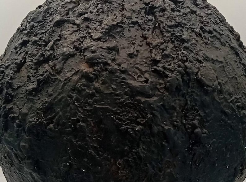 Op de afbeelding: Een bolvormig object met een donkerzwart, sterk getextureerd oppervlak. De buitenkant heeft een ruw, onregelmatig en kraterachtig uiterlijk met diepe groeven en verhoogde bulten, wat een complex, multidimensionaal en tactiel visueel effect cre&euml;ert.