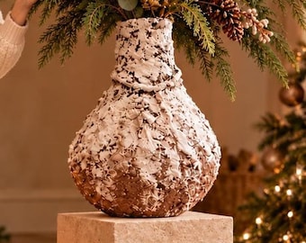 May include: A bulbous ceramic vase featuring a textured white upper section with thick, raised paint strokes and a metallic bronze lower section. The vase is filled with a lush arrangement of green pine branches, eucalyptus leaves, and brown pinecones.