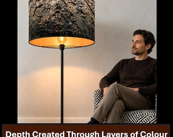 May include: A floor lamp with a tall, thin black metal stand and a cylindrical lampshade featuring a dark, heavily textured, cratered surface. The interior of the shade is a warm, metallic gold, casting a soft glow from the light bulb inside.