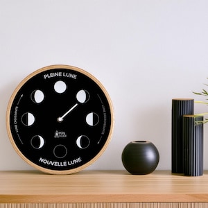 May include: A black and white moon phase clock with a wooden frame. The clock face shows the different phases of the moon, with the words "Pleine Lune" and "Nouvelle Lune" at the top and bottom.