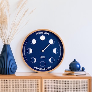 Può includere: Un orologio a fasi lunari blu e in legno con il testo "PLEINE LUNE", "LUNE CROISSANTE", "NOUVELLE LUNE" e "LUNE DECROISSANTE" sul quadrante. L'orologio è su uno scaffale di legno con un vaso blu e libri.