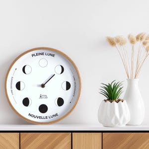 Peut inclure: Une horloge murale ronde avec un cadre en bois et un cadran blanc affichant les phases de la lune. L'horloge a des aiguilles noires et du texte en français. Une petite plante succulente dans un pot géométrique blanc et un vase blanc avec des fleurs séchées sont à côté.