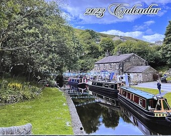 May include: A 2023 calendar featuring a scenic canal view with several black and red canal boats. The image includes a stone building, lush greenery, and a blue sky with clouds. The calendar title is in an elegant script.