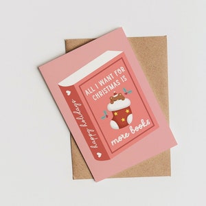 May include: A pink greeting card with a book design and the text "All I want for Christmas is more books." The card features a Christmas stocking illustration with a bear and holly. A brown envelope is behind the card.
