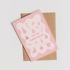 May include: A pink Christmas card with the phrase "'Tis the Damn Season" and festive illustrations of a candy cane, mittens, a Christmas tree, a bell, a star, a stocking, a present, and a mug. The card is placed on a brown envelope.