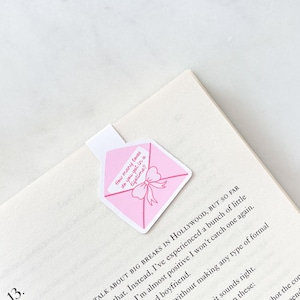 May include: Pink envelope-shaped bookmark with a bow graphic and the text "How many loves do you get in a lifetime?" The bookmark is placed on an open book, enhancing the reading experience.