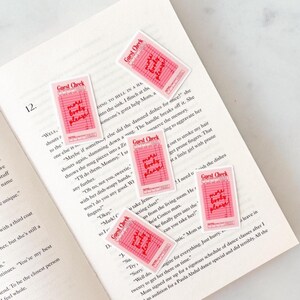 Op de afbeelding: Vier rechthoekige stickers ontworpen als roze gastenbonnen. Elke sticker bevat de tekst "Guest Check" bovenaan en de handgeschreven zin "more books please!" in rode inkt over de rasterlijnen van het papieren formulier heen.