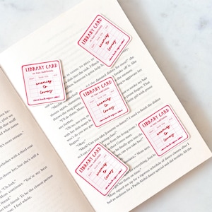 Op de afbeelding: Vijf rechthoekige stickers ontworpen als vintage bibliotheekkaarten met rode randen en tekst. Elke sticker bevat de woorden LIBRARY CARD, on loan indefinitely en de handgeschreven tekst enemies to lovers in rode inkt.