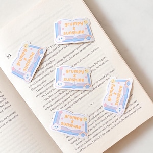 May include: Five die-cut stickers shaped like open books with light blue covers and pages. Each sticker features the orange text "grumpy x sunshine" with a small yellow sun icon, a pink star, and a small blue cloud with a grumpy face at the bottom.