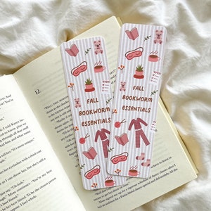 Puede incluir: Dos marcapáginas rectangulares con un patrón de rayas verticales y el texto "FALL BOOKWORM essentials". Los marcapáginas presentan ilustraciones de libros, osos de peluche, tazas de café y antifaces.