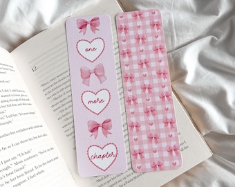 May include: Two pink bookmarks with bow and heart designs rest on an open book. One bookmark features the words "one", "more", and "chapter" within hearts. The other has a pink gingham pattern with bows.