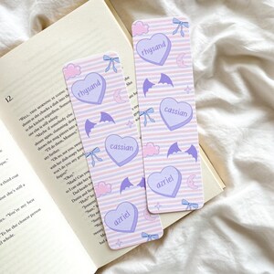 Puede incluir: Dos marcapáginas rectangulares con un fondo a rayas rosas y lilas. Cada marcapáginas presenta diseños en forma de corazón con nombres como "rhysand", "cassian" y "azriel", junto con alas de murciélago, lazos y medias lunas.