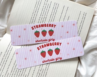 May include: Two rectangular bookmarks with a pink and white striped design. Each bookmark has the word "STRAWBERRY" above three red strawberries and the words "shortcake girly". Small pink flowers are scattered.