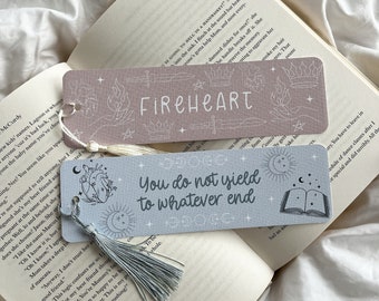 May include: Two rectangular bookmarks with tassels rest on an open book. One bookmark is light pink with the word "FIREHEART" and line art. The other is light blue with the text "You do not yield to whatever end".
