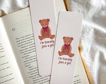 May include: Two rectangular bookmarks with a teddy bear illustration and the text "I'm literally just a girl." The bear is brown with a red bow. The bookmarks are on a book with white pages.