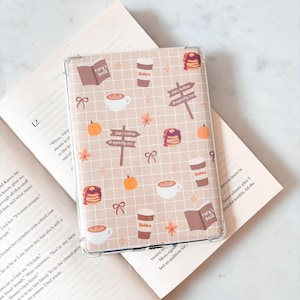 以下が含まれることがあります： ベージュの格子模様にコーヒーカップ、カボチャ、パンケーキ、「Rory's book」と「Stars Hollow Dragonfly Inn」の文字が描かれた、透明な電子書籍リーダー用保護ケース。