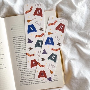 Peut inclure: Deux marque-pages décoratifs sur fond crème, avec des motifs répétés d'écharpe, de pull avec un "H", de lunettes, d'un balai, d'une grenouille et d'un chapeau. Un marque-page a un pull bleu avec un "R". L'autre a un pull bordeaux avec un "H".