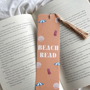 May include: A peach-coloured bookmark with the words "BEACH READ" in white. The bookmark features illustrations of seashells, ice lollies, and beach umbrellas. A tassel hangs from the top. The bookmark is placed in an open book.