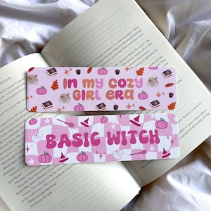 Könnte beinhalten: Zwei rechteckige Lesezeichen mit rosa und weißen Designs. Eines zeigt "IN MY COZY GIRL ERA" mit Kürbissen und Blättern. Das andere zeigt "BASIC WITCH" mit Hexenhüten und Geistern. Beide liegen auf einem offenen Buch.