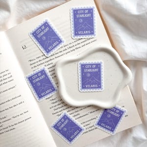 Può includere: Una collezione di adesivi viola con un design a francobollo. Ogni adesivo presenta il testo "CITY OF STARLIGHT" e "VELARIS" con una grafica di montagna e luna. Gli adesivi sono disposti su un libro aperto e un piatto di ceramica bianco.