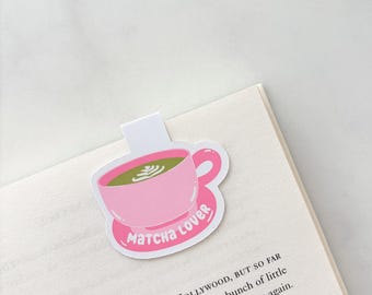 May include: A pink bookmark with a matcha-themed design. The bookmark features a pink teacup filled with green matcha and the words "Matcha Lover". The bookmark is placed on the corner of a book.