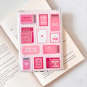 Peut inclure: Coque de téléphone transparente avec un motif à rayures roses et blanches et des autocollants roses. Les autocollants affichent des phrases comme "enemies to lovers" et "hot girls read". La coque est posée sur un livre ouvert.
