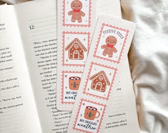 May include: Two festive bookmarks with gingerbread men, gingerbread houses, and hot chocolate mugs. The bookmarks have the text "FESTIVE FEELS" and "HOT CHOCOLATE weather" printed on them. The bookmarks are on top of an open book.