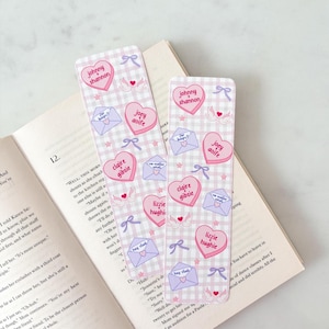 Puede incluir: Dos marcapáginas decorativos con un patrón de cuadros rosa y blanco. Cada marcapáginas presenta diseños en forma de corazón con nombres, junto con ilustraciones de sobres y lazos. Los marcapáginas están sobre un libro abierto.
