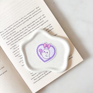 May include: A white, speckled, egg-shaped ceramic dish holds a sticker of a cartoon ghost with a lollipop and a pink bow, inside a purple heart. The dish rests on an open book with text.