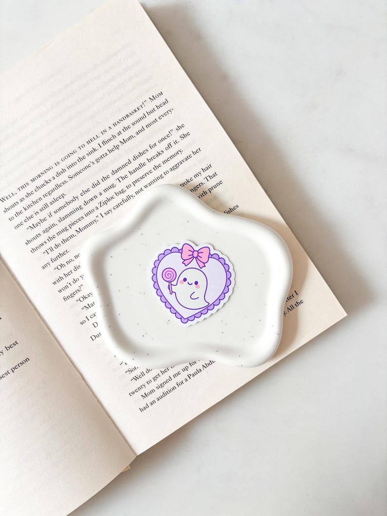 May include: A white, speckled, egg-shaped ceramic dish holds a sticker of a cartoon ghost with a lollipop and a pink bow, inside a purple heart. The dish rests on an open book with text.