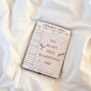 Könnte beinhalten: Transparente Handyhülle mit einem Bibliothekskarten-Design. Das Design enthält die Wörter "LIBRARY CARD", "YOU DO NOT YIELD TO WHATEVER END" und "READ MORE BOOKS". Die Hülle hat ein hellrosa und weißes Farbschema.