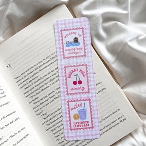 May include: A rectangular bookmark with a pink and white gingham pattern. It features three postage stamp-style designs with text and illustrations: "Welcome to Waning Bay, Michigan," "Cherry Hill Winery," and "Lavender Lemonade."
