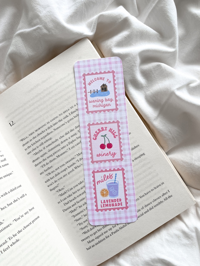 May include: A rectangular bookmark with a pink and white gingham pattern. It features three postage stamp-style designs with text and illustrations: "Welcome to Waning Bay, Michigan," "Cherry Hill Winery," and "Lavender Lemonade."