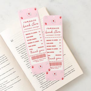 Op de afbeelding: Roze papieren bladwijzers met een "romance book store" ontwerp. De bladwijzers hebben een ontwerp van een klantbon met tekst als "Enemies to Lovers" en "Thank You". Versierd met strikken en aardbeien, liggen ze op een open boek.