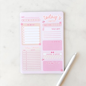 Könnte beinhalten: Ein rosa-weißes Agenda-Notizblock mit den Worten "today's agenda" und Abschnitten für To-Do-Listen, Ziele und tägliche Affirmationen. Ein weißer Stift liegt daneben.