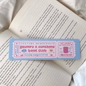 May include: A light blue bookmark with a pink ticket design. The bookmark reads "GRUMPY X SUNSHINE BOOK CLUB" with a cute sun and cloud graphic. The text "he hates everyone but her" is also included. The bookmark is placed on top of an open book.