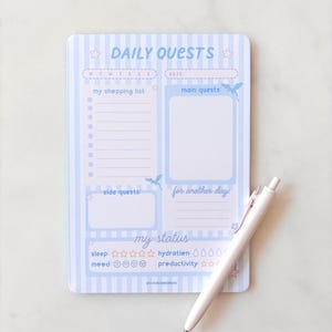 May include: A light blue and white striped daily planner notepad with the text Daily Quests, featuring sections for a shopping list, main quests, side quests, and status trackers for sleep, hydration, mood, and productivity, accompanied by a white pen.