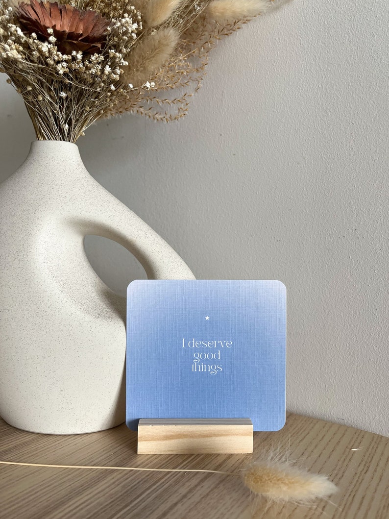 Puede incluir: Una tarjeta azul claro con el texto "I deserve good things" en un bloque de madera. Un jarr&oacute;n blanco con una forma &uacute;nica y flores secas en el fondo.